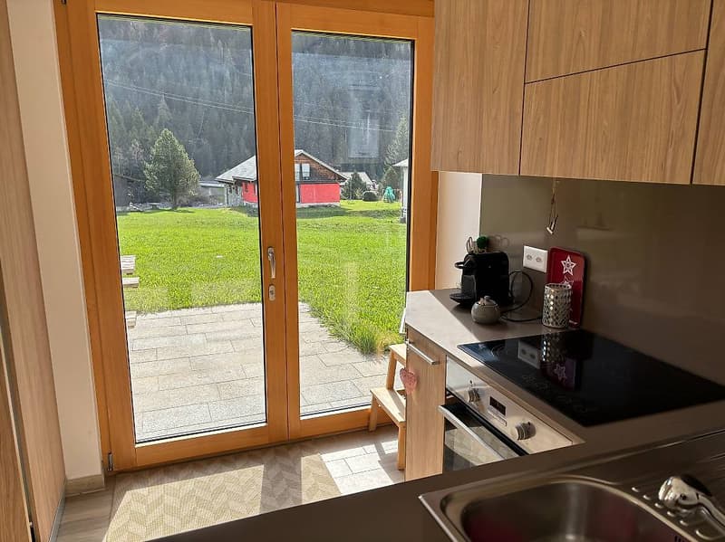Modern kitchen with sliding doors to a terrace, granite countertops, wooden cabinets, black induction stove, oven, wooden stool, grassy view