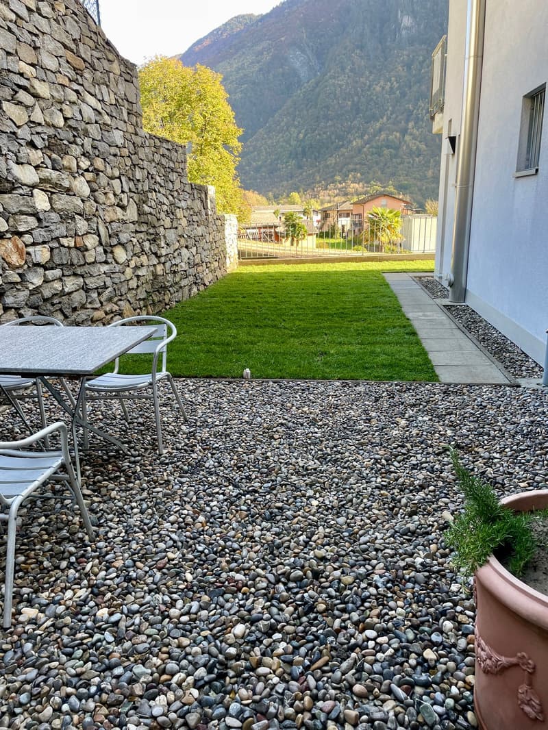 Pebbled patio, table with chairs, potted plant, stone wall, fenced lawn, mountain view