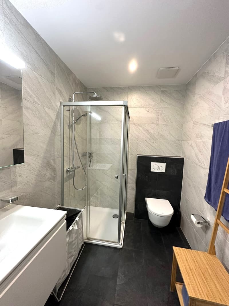 Spacious bathroom with gray tile walls, glass shower enclosure, vanity with drawers, and a toilet with a black accent wall.