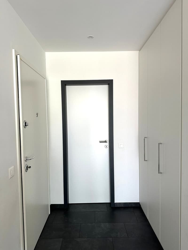 The image shows a hallway with a white wall, a black framed door, and a black tile floor. There is a white door on the left side of the hallway.