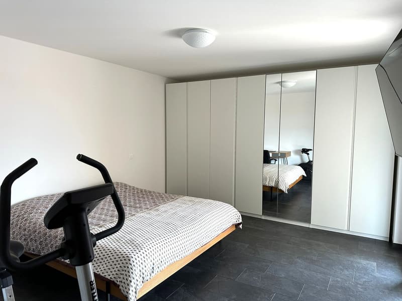 Spacious bedroom with white walls, large mirrored closet, dark tile flooring, and a patterned bedspread on the bed. There is also a piece of exercise equipment visible in the room.