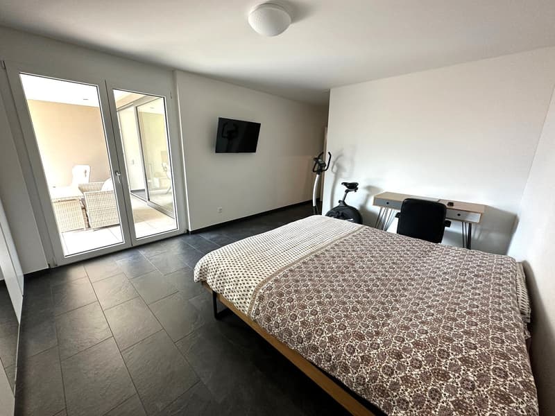 Spacious bedroom with large windows, tile flooring, a double bed with patterned bedding, a TV mounted on the wall, and exercise equipment in the corner.