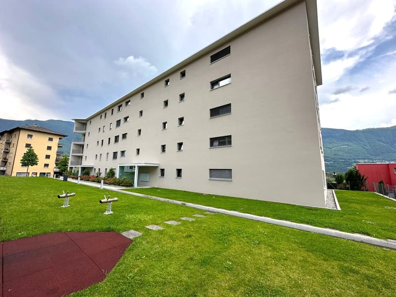 Multi-story apartment building with beige exterior, large windows, and a grassy area with playground equipment in the foreground.