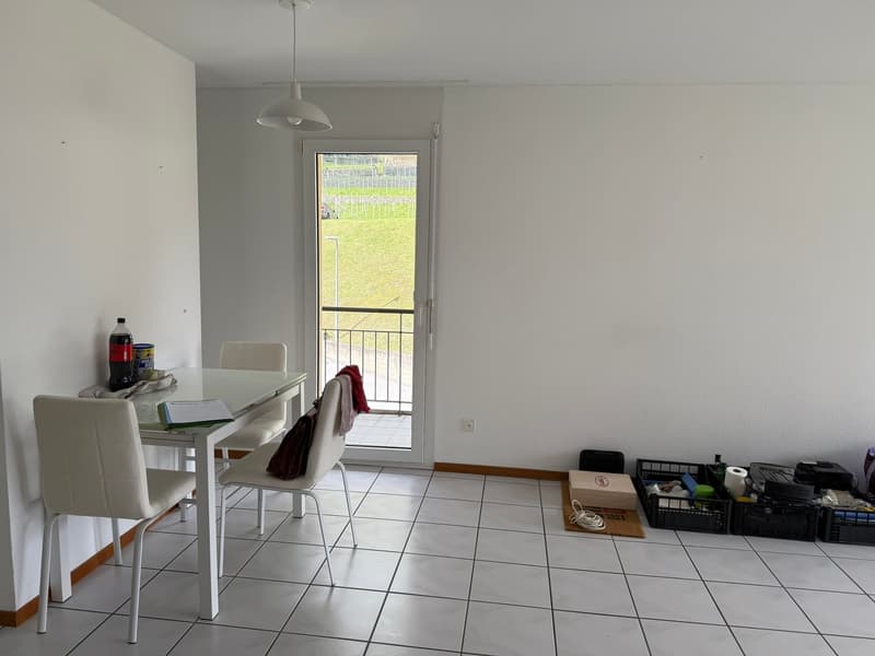 Bright and spacious living room with a dining area, tiled flooring, and a balcony or terrace visible through the window. The room contains a dining table with chairs, a small desk, and some storage boxes and items.