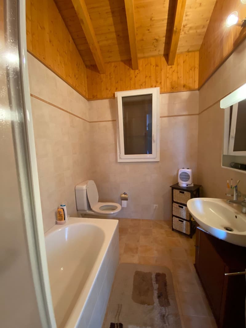 Bathroom with wood-paneled ceiling, tiled walls and floor, bathtub, toilet, vanity with sink, and a small storage unit.