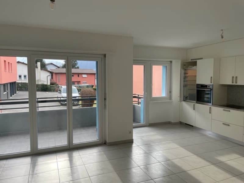 Spacious open-plan living area with large windows and sliding glass doors leading to a balcony or terrace, modern kitchen with white cabinets and appliances, tiled flooring throughout