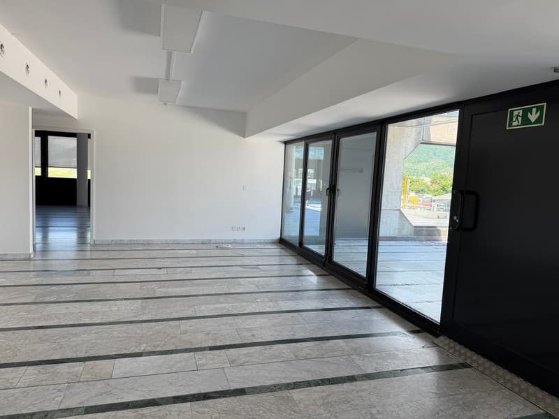 Large open space with white walls, gray tile flooring, and floor-to-ceiling glass doors leading to a balcony or terrace with a view of the outdoors.