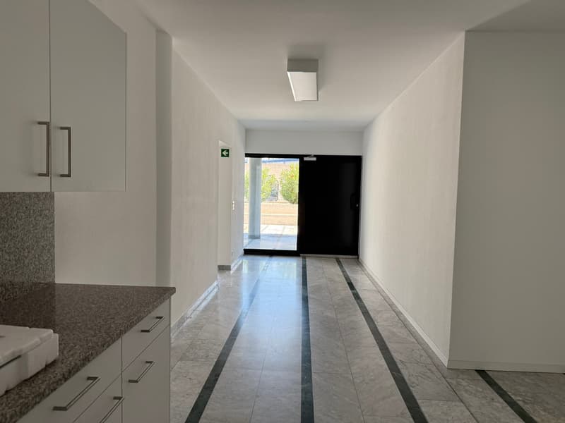 The image shows a long, narrow hallway with white walls and a tiled floor. The hallway has built-in cabinets or storage units along one side, and a doorway at the end leading to an outdoor area. The lighting appears to be recessed and the overall aestheti