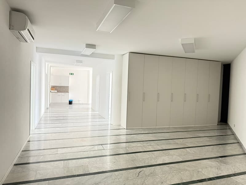 The image shows a large, open office or commercial space with a long, wide hallway. The hallway has a sleek, modern design with white walls, gray tile flooring with black accents, and recessed lighting fixtures in the ceiling. At the end of the hallway, t