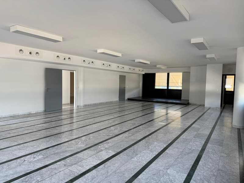 Large open space with gray marble-like flooring, white walls, and recessed lighting fixtures. The space appears to be a commercial or office area, with a door visible in the background.