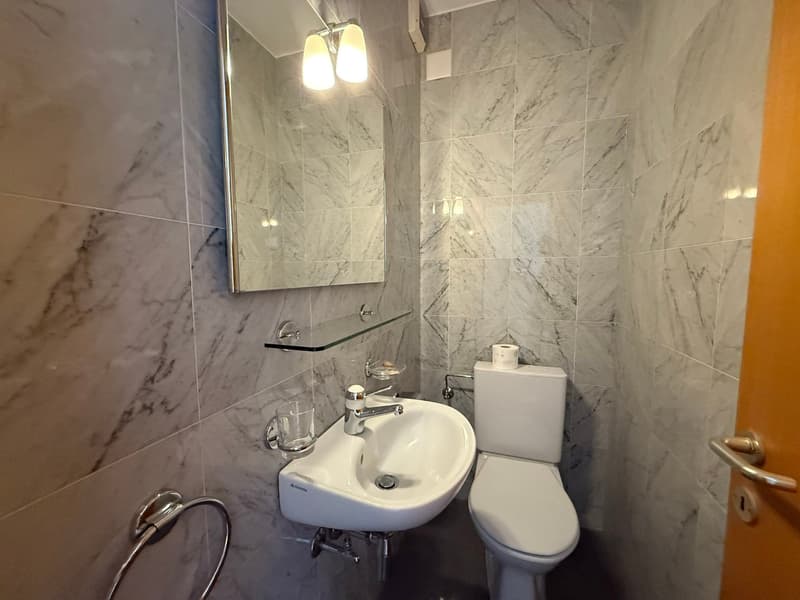 The image shows a bathroom with marble-like tile walls, a small sink with a mirror above it, and a toilet. The bathroom appears to be well-lit with a pair of light fixtures on the wall.