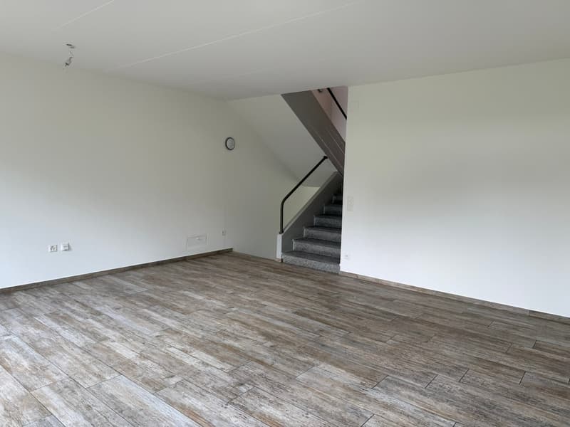 The image shows an empty room with a hardwood-style floor and white walls. There is a staircase visible in the corner of the room.