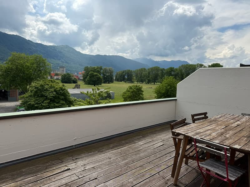The image shows an outdoor terrace or balcony overlooking a scenic landscape. The terrace has a wooden floor and some wooden furniture, including a table and chairs. The view from the terrace includes a grassy field, trees, and mountains in the background