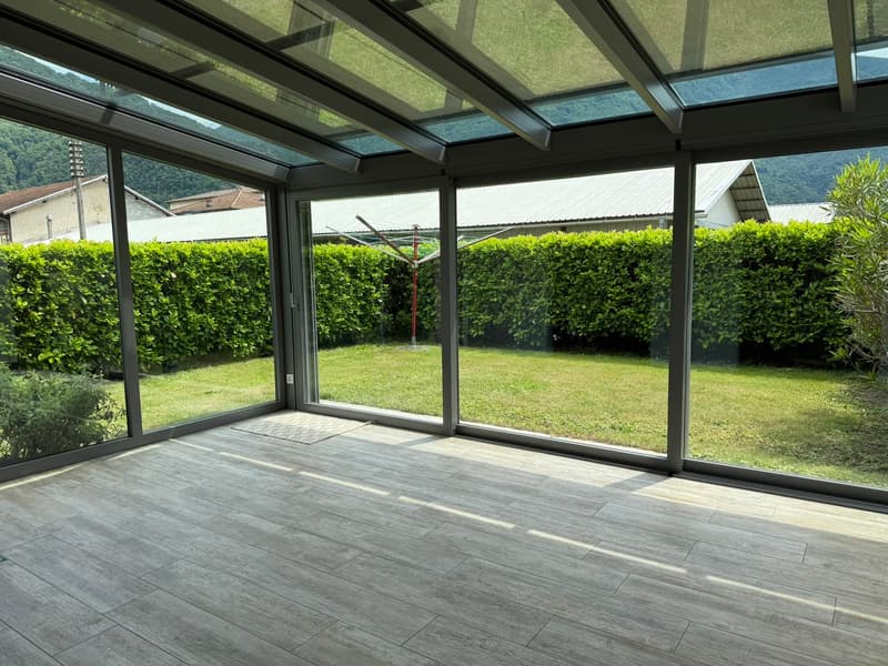 Spacious sunroom with glass ceiling and walls, providing a panoramic view of the lush green garden outside. The room features a wooden floor and a cozy, open layout.