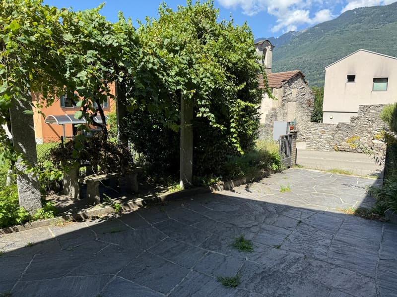 The image shows an outdoor area with a paved stone floor, surrounded by lush green trees and vegetation. There appears to be a balcony or terrace visible on one of the buildings in the background.