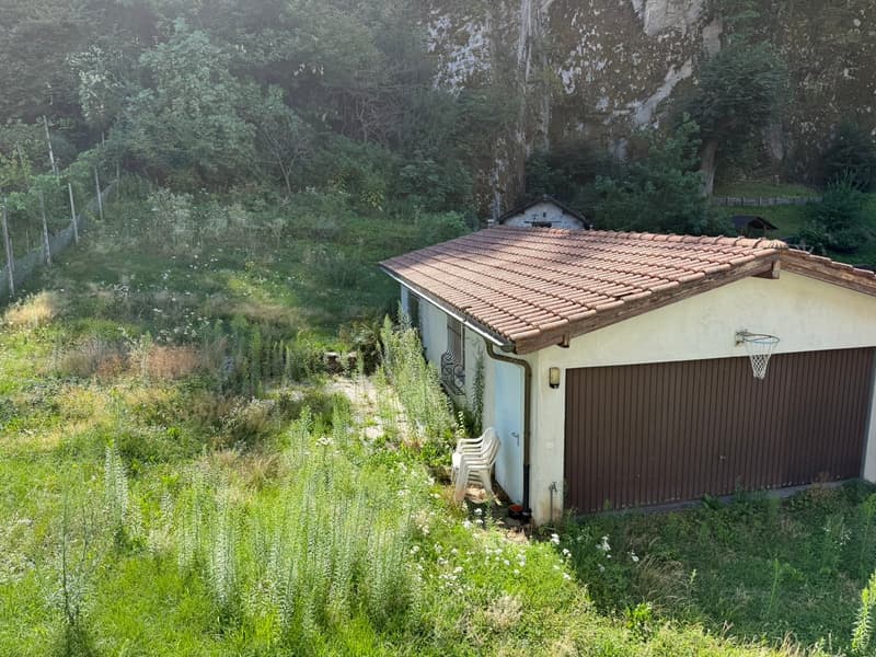 A small house with a tiled roof, surrounded by lush greenery and vegetation. There appears to be a garage or outbuilding attached to the side of the house.
