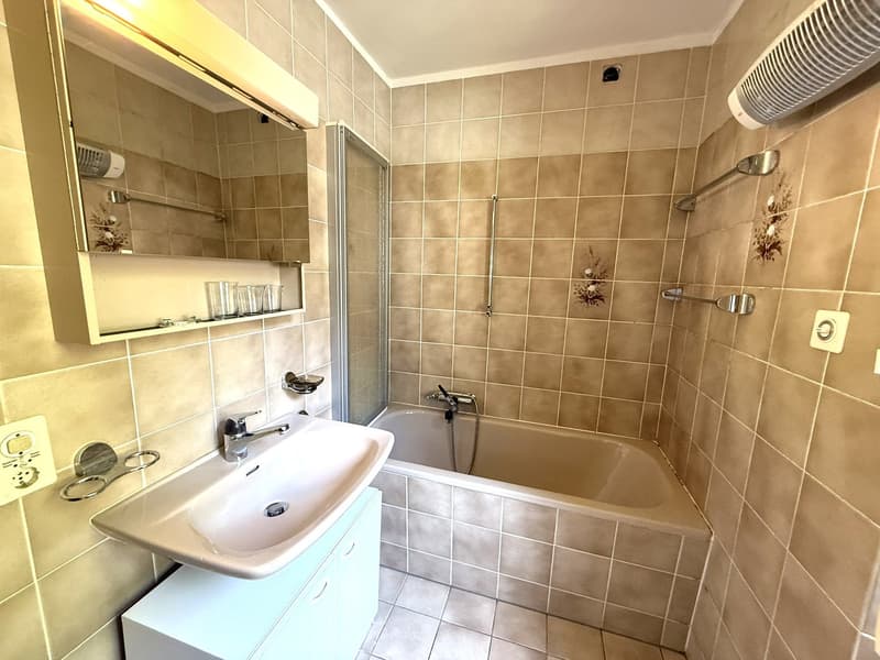 Tiled bathroom with a bathtub, sink, and storage shelves. The bathroom has a neutral color scheme with beige tiles and a white sink.