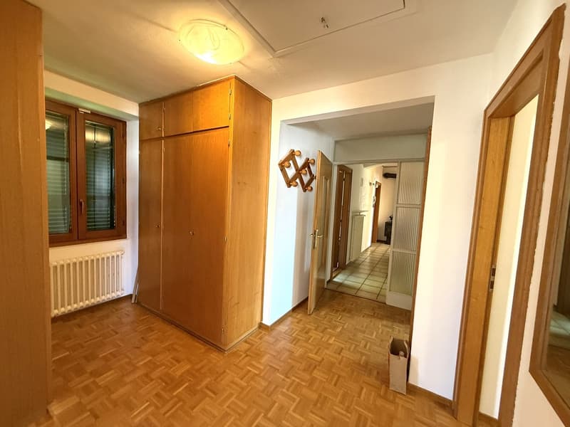The image shows the interior of an apartment. The room has wooden floors and a large wooden cabinet or wardrobe. There is a hallway visible in the background, leading to other rooms. The lighting appears to be warm and the overall atmosphere is cozy.