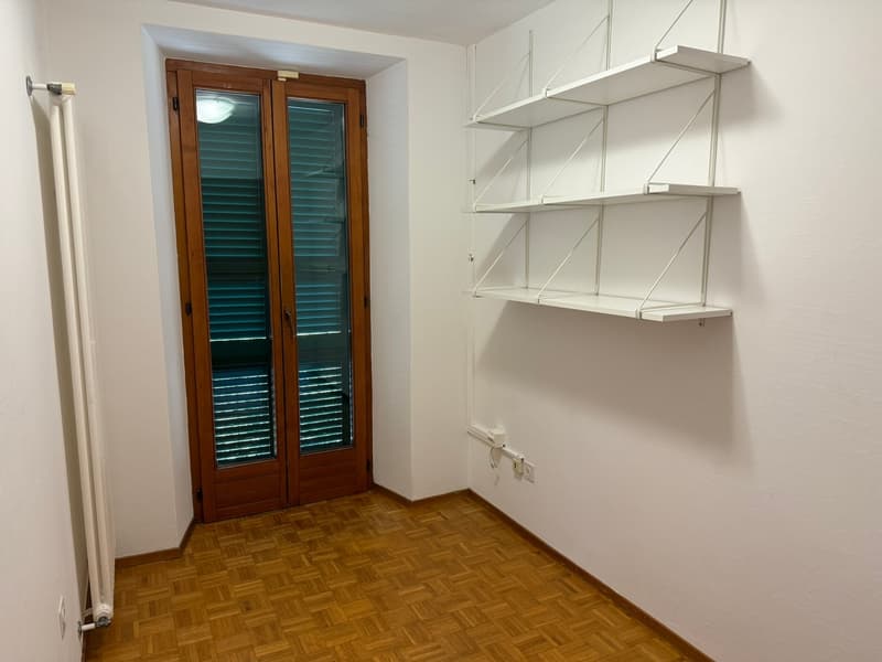 The image shows a small room with white walls, a wooden floor, and a set of wooden shuttered windows. There is a metal shelving unit along one wall, providing storage space. The room appears to be a utility or storage space, with a simple and functional d