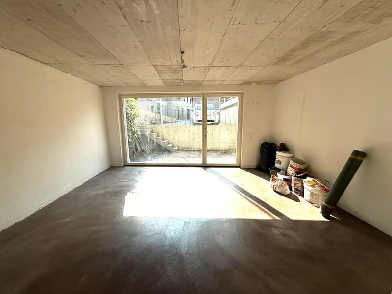 Ample room with sliding glass door, exposed concrete walls and floor, storage containers and a green cylindrical item
