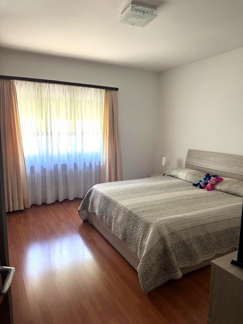 Bedroom with wooden floor, large bed, nightstand, lamp, and a window with curtains.