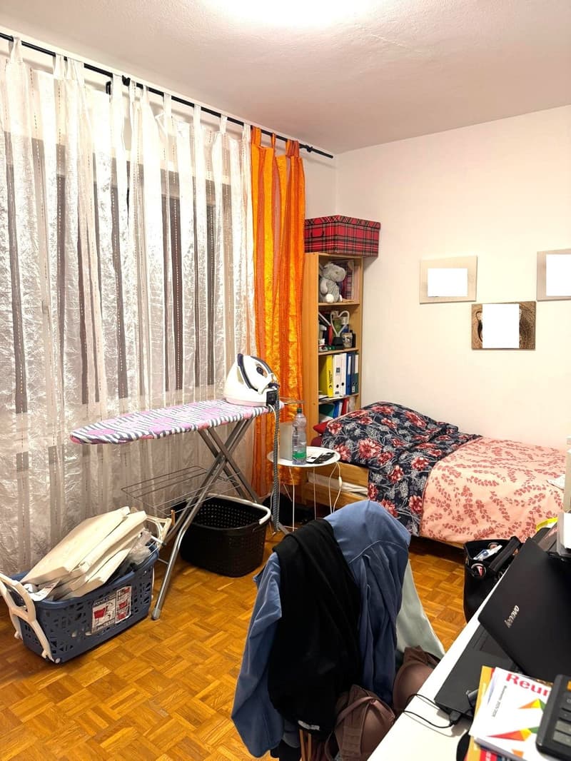 single bed, desk, shelves, iron and ironing board, baskets, table with a bottle and cell phone, curtains