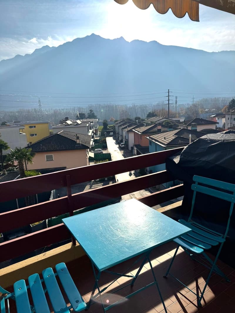 Balcony with wooden railing, blue chairs, square table, covered grill, buildings and mountains in the distance