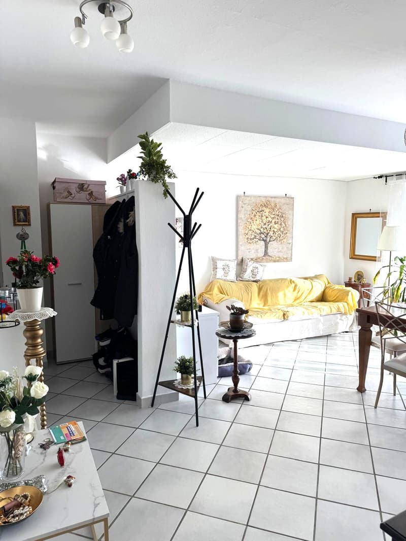 open space living room with dining area, light white tiles, white ceiling, three lights, sofa, couch, coat rack, stand, side table, chairs, dining table, small white cabinet