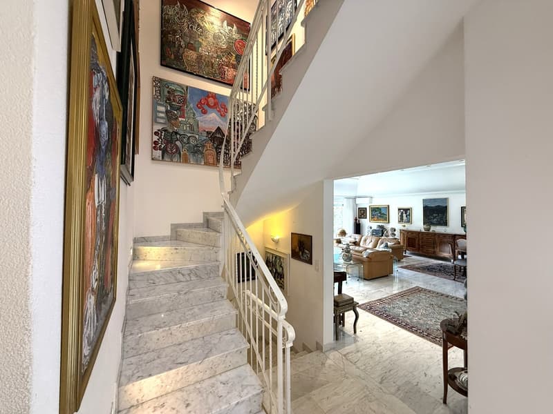 The hallway features marble stairs, white railings, a few paintings on the wall, and a beige carpet at the bottom of the stairs.