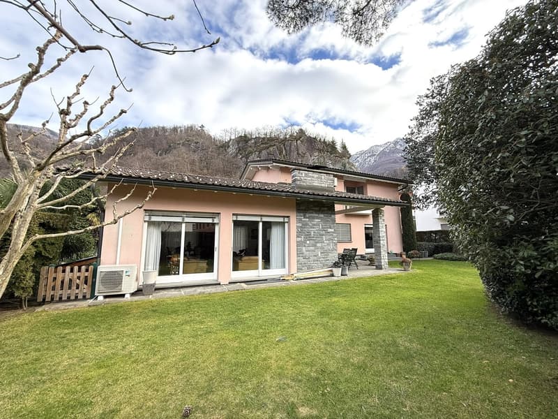 Detached house, pink exterior, large windows, spacious lawn, exterior AC unit, patio with chairs, trees, bushes, mountain in the background