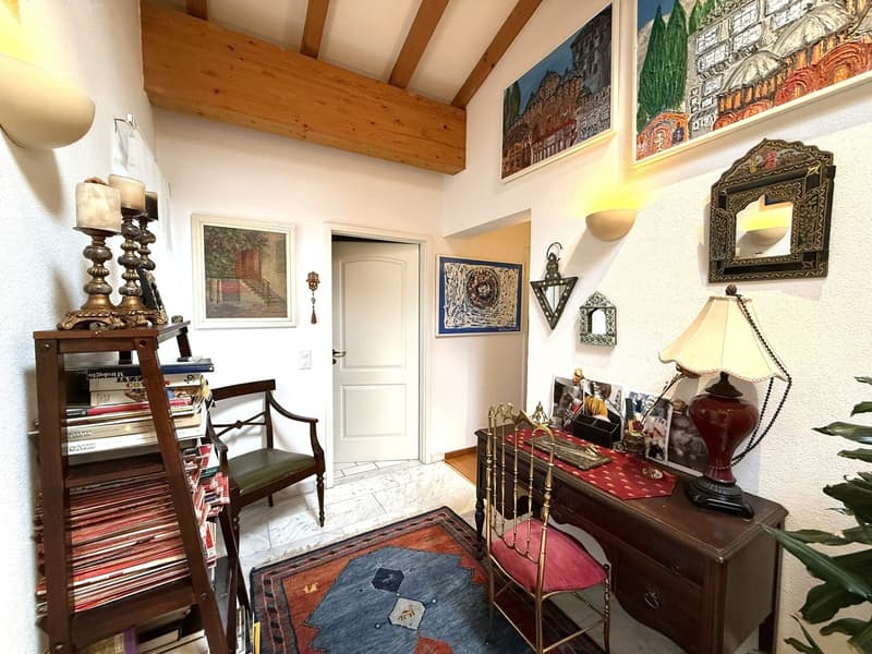 wooden beams on ceiling, decorative chair, white wall, books on shelf, carpet, framed pictures on wall, wooden desk, red cloth on desk, lamp on desk, photo frames on desk
