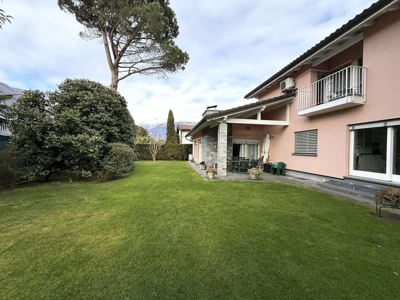 Two-story pink house, white-framed windows, metal balcony with metal railings, a garden, several bushes, and a large grassy lawn, an outdoor seating area with chairs and a table, a tree