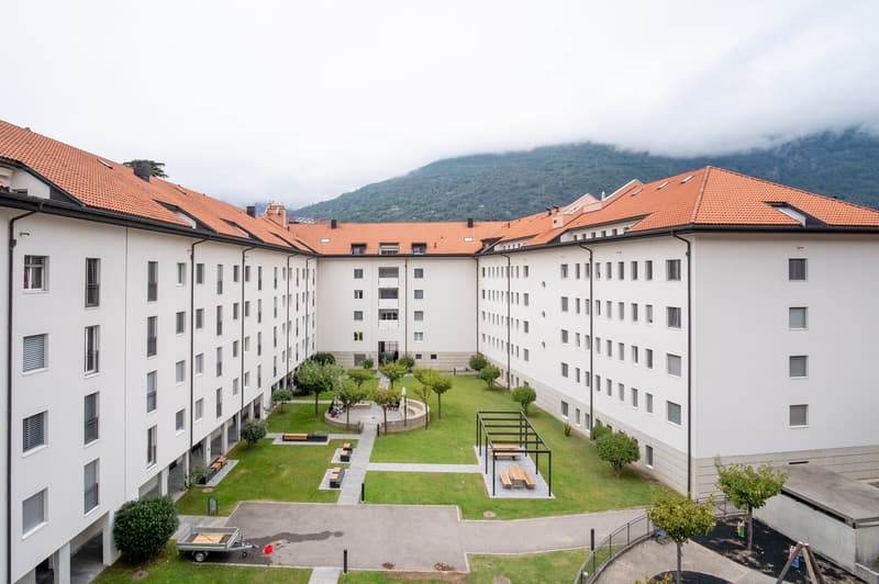 Modern apartment building with multiple floors, red roofs, white walls, central courtyard with grass, benches, and a pergola, surrounded by trees and plants, with a mountain in the background.