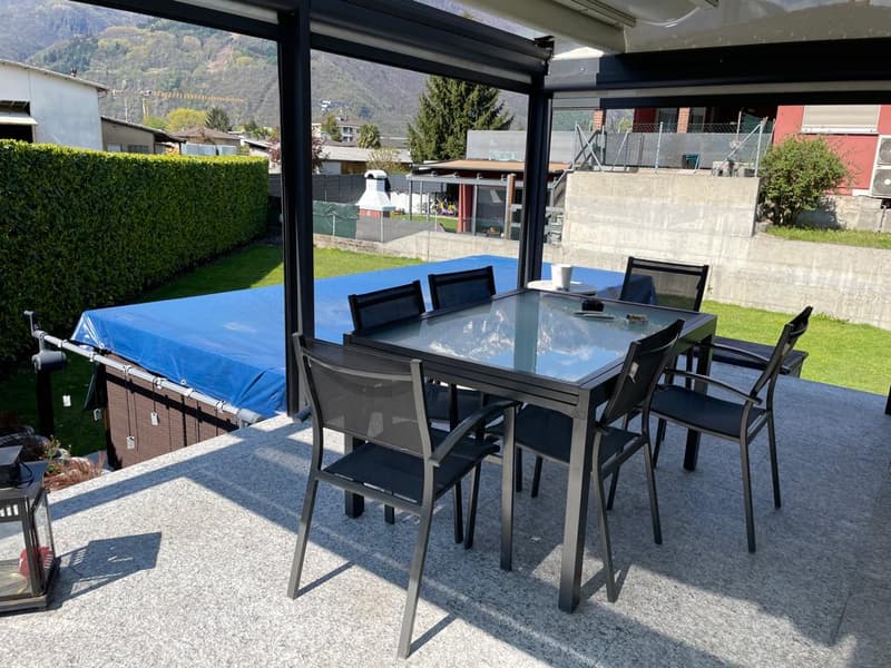 Outdoor dining table with chairs, covered by a retractable roof, surrounded by a garden with a hedge and a grassy lawn, with a view of the mountains