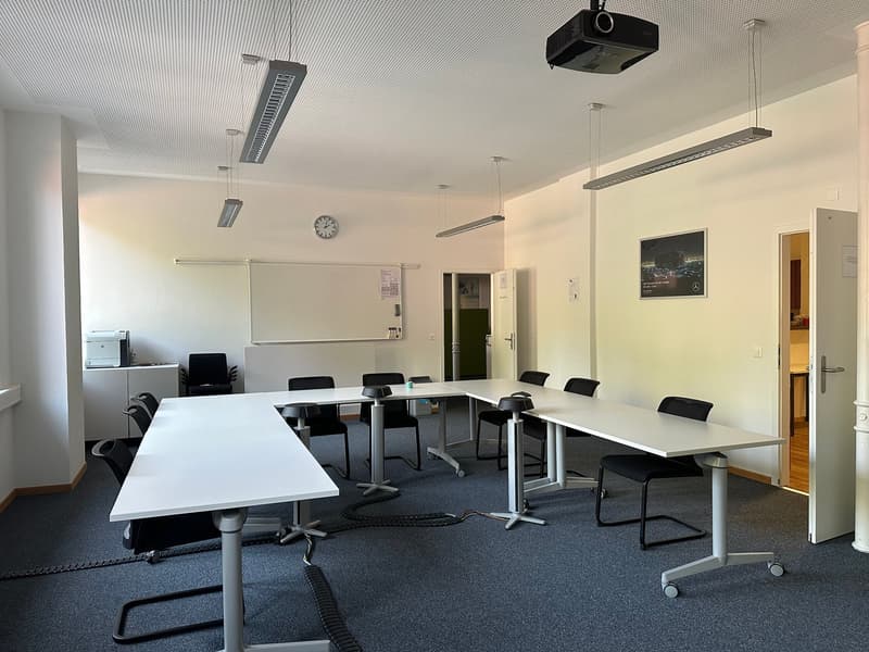 The image shows a modern office space with a large conference table surrounded by black chairs. The room has white walls, a projector, and several hanging light fixtures. There is also a clock on the wall and a framed picture. The floor is covered in a da