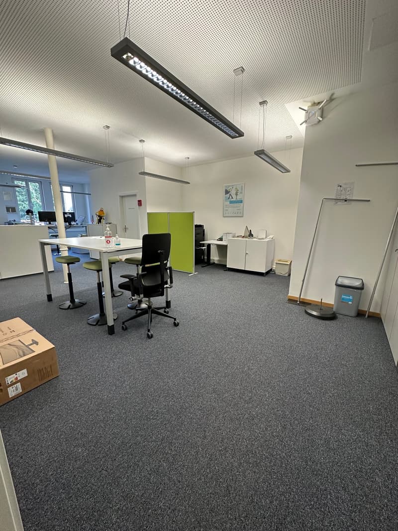 The image shows an office space with a modern and minimalist design. The room has a gray carpet floor, white walls, and suspended linear lighting fixtures. There are several desks with office chairs, a printer, and some storage units. The overall layout a