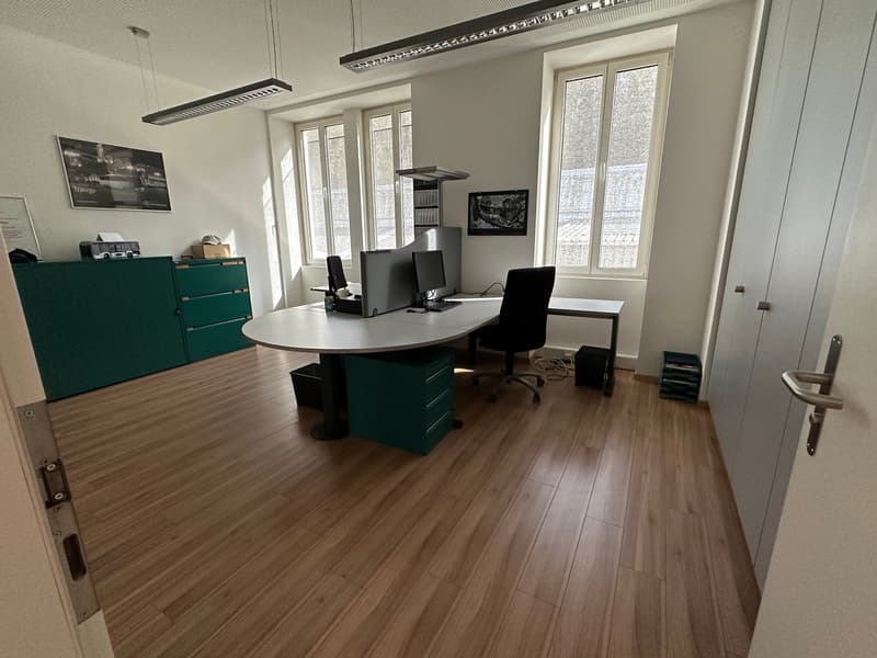 The image shows a modern office space with a round desk, computer setup, and storage cabinets. The room has large windows, hardwood flooring, and a minimalist, professional decor.
