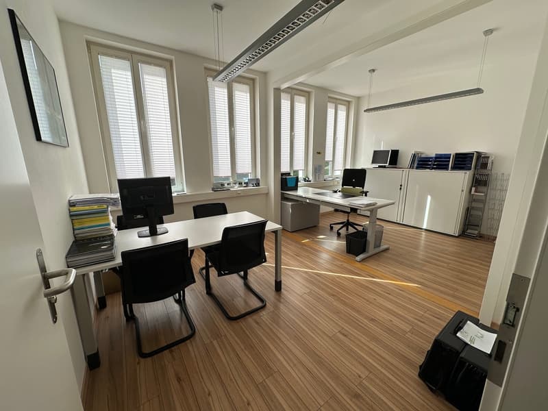 Spacious office with large windows, hardwood floors, desks, computers, and storage units