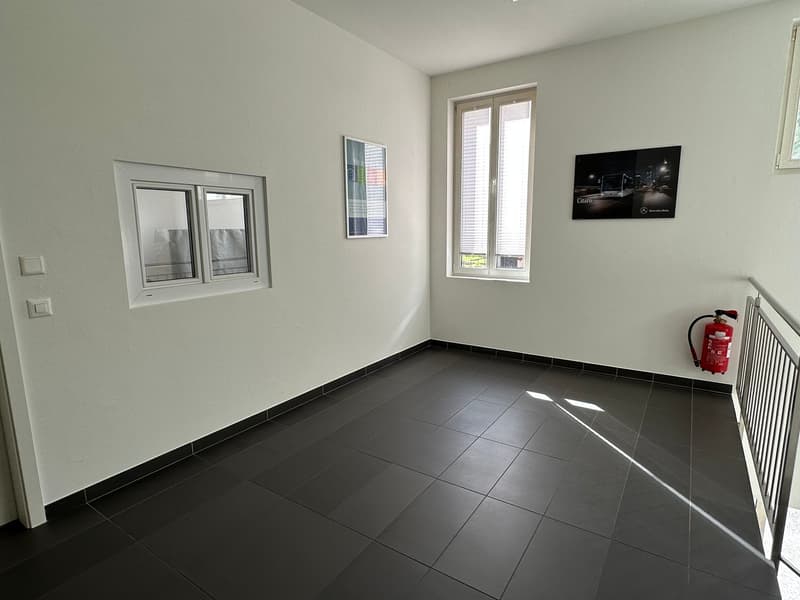 The image shows a room with white walls, a large window, and a black and white framed artwork on the wall. The floor is covered with dark gray tiles. There is a fire extinguisher visible in the corner.