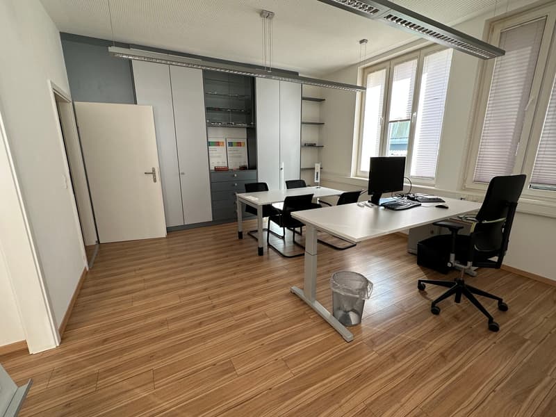 Spacious office with hardwood flooring, white walls, adjustable desks, office chairs, and built-in storage cabinets. There is a computer setup on one of the desks, indicating this is a workspace or office environment.
