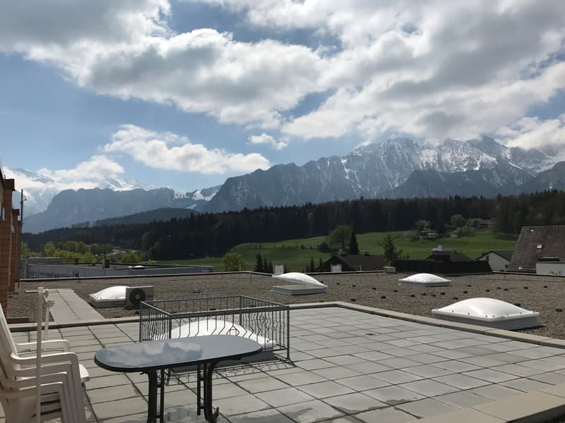rooftop terrace, chairs, table, fenced balcony, snowy mountains, cloudy sky, green hills, buildings, trees