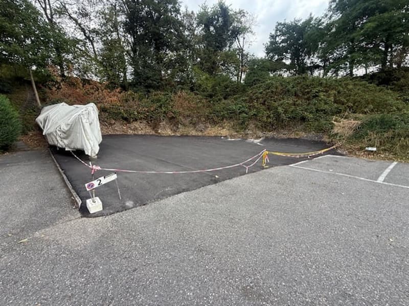 Outdoor parking area with a covered vehicle, marked with caution tape and numbered signs, surrounded by greenery and trees.