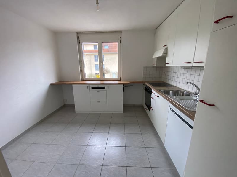 Empty kitchen with white cabinets, tiled floor, and a window overlooking a building.