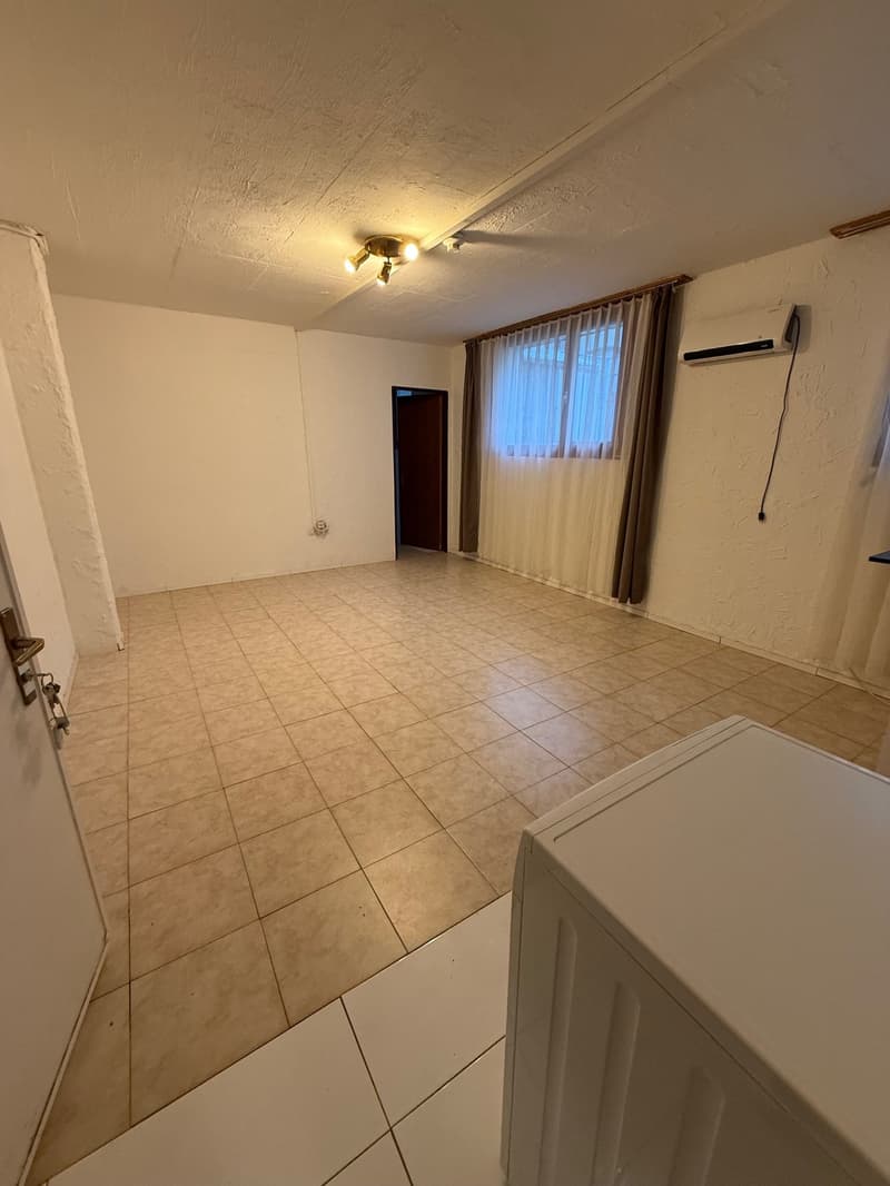 an empty room with tiled floor, white walls, a ceiling with lights, a window with white and brown curtains, a door, and an air conditioner on the wall