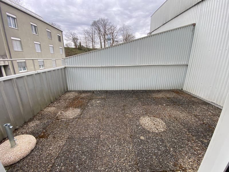 Gravel-covered balcony area enclosed by a metal fence, with a multi-story building in the background and a small circular platform with a pipe.