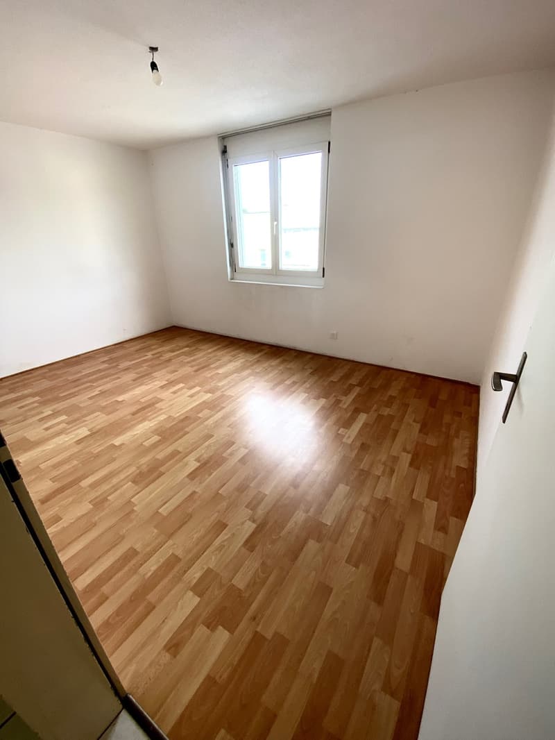 Spacious bedroom with hardwood flooring, white walls, and a single window providing natural light.
