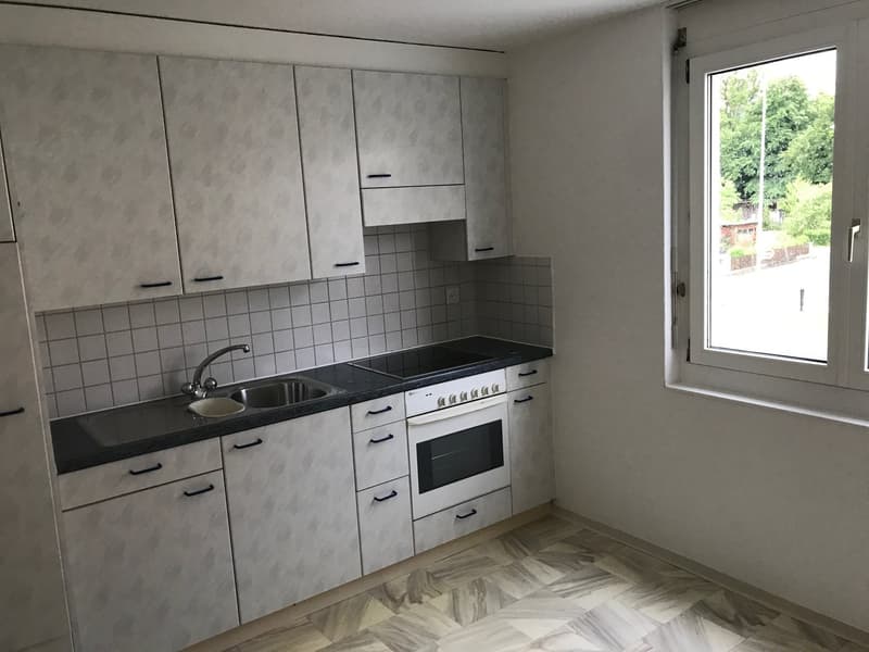 Modern kitchen with gray cabinets, black countertop, built-in oven and stove, sink, and tiled backsplash. Large window providing natural light.