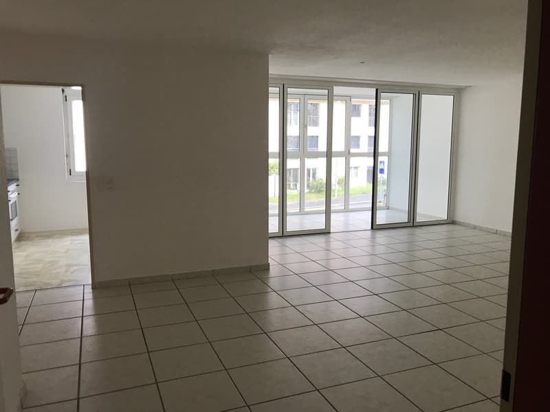 The image shows a spacious apartment with large windows, a tiled floor, and a minimalist interior design. The room appears to be a living area or open-plan living space, with a view of the outdoors visible through the windows.