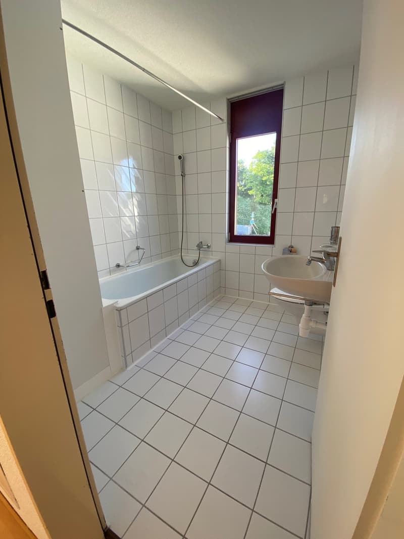 Modern bathroom with white tiled walls and floor, featuring a bathtub, sink, and toilet. A window with a purple frame provides natural light.