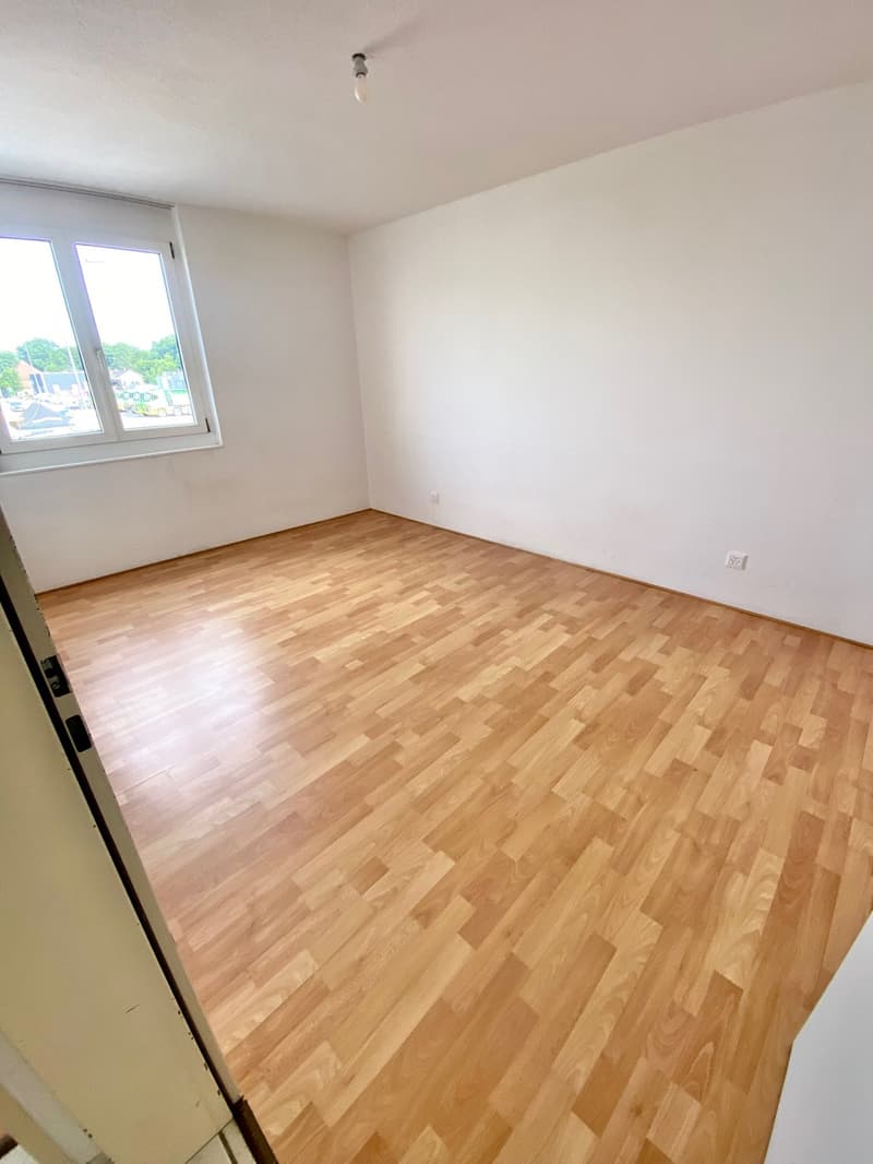 Spacious bedroom with hardwood flooring, white walls, and a large window providing natural light.